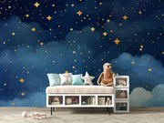 Starry night wall mural with golden stars and deep blue watercolor-style clouds, decorating a cozy bedroom with patterned bedding, soft toys and storage.