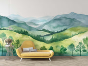 Watercolor green hills wall mural featuring lush rolling landscapes and soft blue mountains, styled in a cozy modern living room with a yellow sofa.