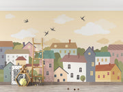 Whimsical village wall mural featuring pastel houses, trees, clouds, and flying birds, styled in a cozy and creative kids’ playroom.