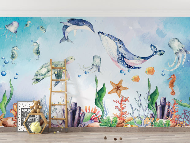 Underwater-themed kids wall mural with watercolor illustrations of a whale, turtle, dolphin, jellyfish, seahorses, coral, and fish in a nursery setting.