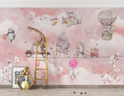 A whimsical nursery wall mural featuring playful animals, including a giraffe on a scooter, an elephant on a bike, and a panda floating with balloons over pink clouds.