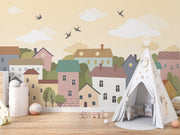 Whimsical village wall mural featuring pastel houses, trees, clouds, and flying birds, styled in a cozy and creative kids’ playroom.