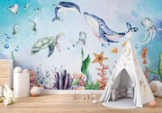 Underwater-themed kids wall mural with watercolor illustrations of a whale, turtle, dolphin, jellyfish, seahorses, coral, and fish in a nursery setting.