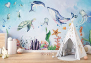 Underwater-themed kids wall mural with watercolor illustrations of a whale, turtle, dolphin, jellyfish, seahorses, coral, and fish in a nursery setting.