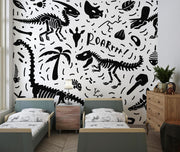 Black-and-white dinosaur-themed wall mural with skeletons, fossils, and "ROAR" text, decorating a kids' bedroom with a cosy bed, playful bedding and a modern lamp.