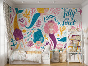Colorful mermaid-themed wall mural in a children's playroom, featuring smiling cartoon mermaids with vibrant tails, sea creatures and coral.
