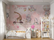 A whimsical nursery wall mural featuring playful animals, including a giraffe on a scooter, an elephant on a bike, and a panda floating with balloons over pink clouds.