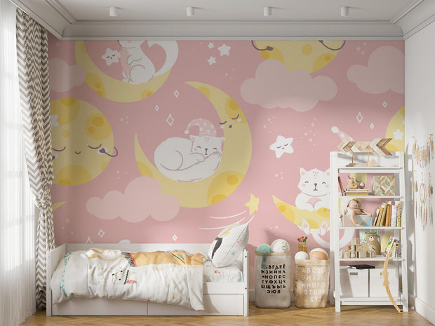 Moon-and-cat-themed wall mural featuring pastel pink tones, crescent moons, sleepy cats and stars, decorating a cozy nursery with soft bedding, toys, and storage shelves.