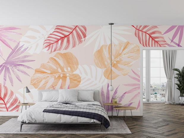 Colorful tropical leaf wall mural featuring overlapping monstera and palm leaves in soft orange, pink, and red tones, styled in a cozy bedroom.