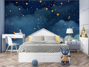 Starry night wall mural with golden stars and deep blue watercolor-style clouds, decorating a cozy bedroom with patterned bedding, soft toys and storage.