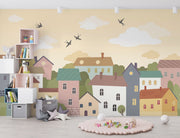 Whimsical village wall mural featuring pastel houses, trees, clouds, and flying birds, styled in a cozy and creative kids’ playroom.
