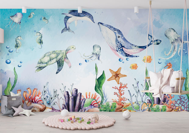 Underwater-themed kids wall mural with watercolor illustrations of a whale, turtle, dolphin, jellyfish, seahorses, coral, and fish in a playroom setting.
