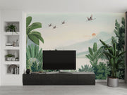 Scenic jungle wall mural with green tropical leaves, misty mountains, and flying cranes against a soft sunrise backdrop.