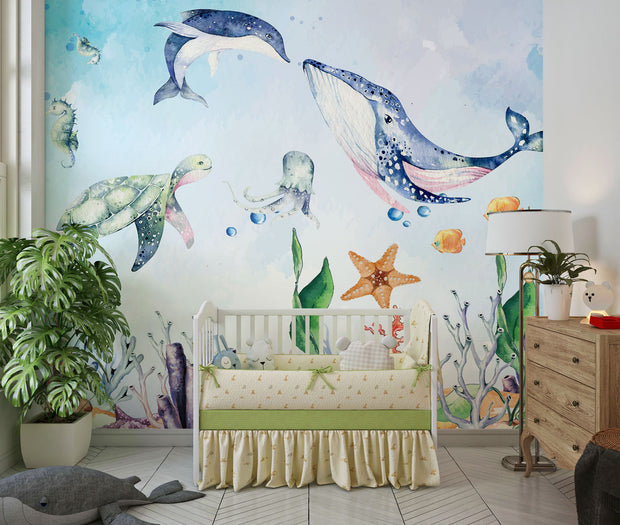 Underwater-themed kids wall mural with watercolor illustrations of a whale, turtle, dolphin, jellyfish, seahorses, coral, and fish in a nursery setting.