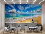 Serene beach wall mural featuring turquoise ocean waves, golden sand and a blue sky with fluffy clouds, decorating a modern office space with stylish furniture.