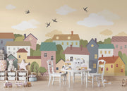 Whimsical village wall mural featuring pastel houses, trees, clouds, and flying birds, styled in a cozy and creative kids’ playroom.