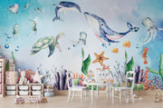 Underwater-themed kids wall mural with watercolor illustrations of a whale, turtle, dolphin, jellyfish, seahorses, coral, and fish in a playroom setting.