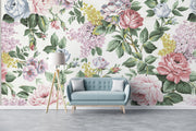 Elegant vintage floral wall mural with pink roses, lilacs, and greenery behind a blue sofa in a cozy, light-filled living room.