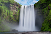 Skógafoss waterfall photograph with white cascades between green cliffs, mist and a pebbled foreground
