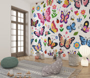 A colorful children's room featuring a wall covered in a whimsical butterfly and flower wallpaper mural. The room is styled with a gray patterned rug, a green knitted pouf, a gray bunny cushion, a white paper lantern lamp, and a wooden French door in the background.