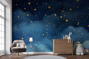 Starry night wall mural with golden stars and deep blue watercolor-style clouds, decorating a cozy bedroom with patterned bedding, soft toys and storage.
