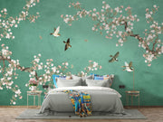 A styled bedroom featuring a large wall mural with a teal-green background, white and pink cherry blossom branches, flying birds, and a bed dressed in gray bedding with a colorful throw blanket.