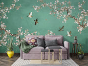 A styled living room featuring a large green wall mural with cherry blossom branches and flying birds, behind a lavender sofa with decorative pillows and a throw blanket.