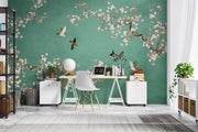 A styled home office featuring a prominent green wall with a delicate cherry blossom branch and bird mural. In front of the wall is a light wood desk with white trestle legs, a white chair, a globe, a laptop, and a desk lamp, set against a gray wood floor.