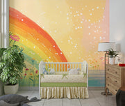 A colorful children's nursery featuring a large wall mural with a rainbow, yellow and peach sky, and white speckles. A white wooden crib with a green and cream bedding set is centered in front of the mural, with a potted plant on the left and a wooden dresser with a lamp on the right.