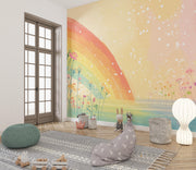 A children's room interior featuring a large wall mural with a pastel rainbow, flowers, and floating white dots on a warm yellow and peach background. The room includes a gray beanbag, patterned rug, green pouf, small decorative figures, and a white floor lamp.