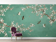 A styled interior scene showing a large wall mural with a teal-green background, white and pink cherry blossom branches, and several flying birds. A purple upholstered armchair with gold legs sits in front of the wall on a light wood floor.
