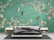 A modern bedroom featuring a large wall mural with a green background, white and pink cherry blossom branches, and several flying birds. A low white platform bed with dark bedding is centered against the wall, flanked by two light wood nightstands with lamps.