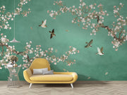 A styled interior scene showing a large wall mural with a teal-green background, white and pink cherry blossom branches, and several flying birds. In front of the wall is a mustard yellow lounge chair with two gray pillows, plus a small wire side table with a hanging lamp nearby.