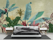 A tropical wallpaper mural featuring large green and teal banana leaves, red heliamericans, and a textured beige background, styled in a modern bedroom setting behind a white platform bed with dark gray bedding.