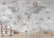 A children's room featuring a large world map wall mural decorated with hot air balloons, ships, airplanes, clouds, and small illustrated landmarks in muted gray, beige, and dusty rose tones. A small white table and chairs, toys, and a storage shelf are arranged in front of the mural.