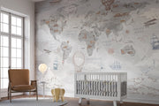 A styled nursery room featuring a large world map wallpaper mural with vintage illustrations of ships, airplanes, hot air balloons, and clouds in muted gray, beige, and brown tones. A white wooden crib, a brown leather armchair, a small side table with a glowing globe lamp, and a round rug are arranged in front of the wall.
