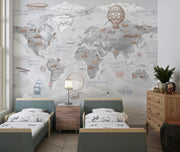 A children's bedroom featuring a large world map wall mural with vintage illustrations of ships, airplanes, and hot air balloons. Two small beds with matching patterned bedding, a wooden nightstand with a lamp, and a wooden dresser with a plant are arranged in front of the mural.