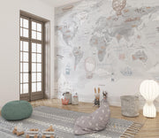 A styled children's room featuring a large world map wall mural with illustrated travel motifs like hot air balloons, ships, and airplanes. The room includes a wooden floor, a patterned rug, a green pouf, a gray bunny cushion, and a white floor lamp.