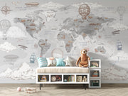 A styled children's room featuring a large world map wall mural with vintage travel illustrations such as hot air balloons, ships, airplanes, and clouds. In front of the wall is a bench with decorative pillows and a plush sloth toy, plus small storage shelves and a light wood floor.