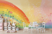 A children's playroom featuring a large, colorful rainbow wall mural with wildflowers and white speckles. The room is styled with a small white table and chairs, storage bins, and plush toys.