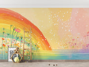 A colorful children's room wall mural featuring a large rainbow arching over a field of wildflowers, with a pastel sky in yellow, pink, and peach tones, blue water at the bottom, and a wooden ladder leaning against the wall.