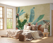 A styled bedroom interior featuring a large tropical wallpaper mural with oversized green banana leaves and red flowers on a warm beige background, behind a bed with neutral-toned bedding.