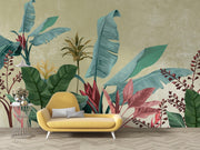 A styled interior scene showing a large tropical wall mural with oversized banana leaves and red foliage on a textured beige background, behind a yellow lounge chair and a small side table.