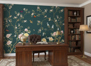A styled home office featuring a dark teal wallpaper with a gold vine pattern, pink and white flowers, and small birds. The room is furnished with a dark wood desk, a matching leather chair, a tall bookshelf, and a floor lamp.