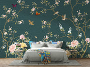 A styled bedroom featuring a large wall mural with a dark teal background, decorated with gold branches, pink and white flowers, butterflies, and birds. A gray bed with white bedding and colorful pillows is positioned in front of the wall.