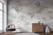 A styled nursery or children's room featuring a large world map wall mural with vintage illustrations of hot air balloons, airplanes, ships, and animals. The room includes a gray upholstered armchair, a wooden dresser, a small side table with a lamp, a round rug, and a plush toy.