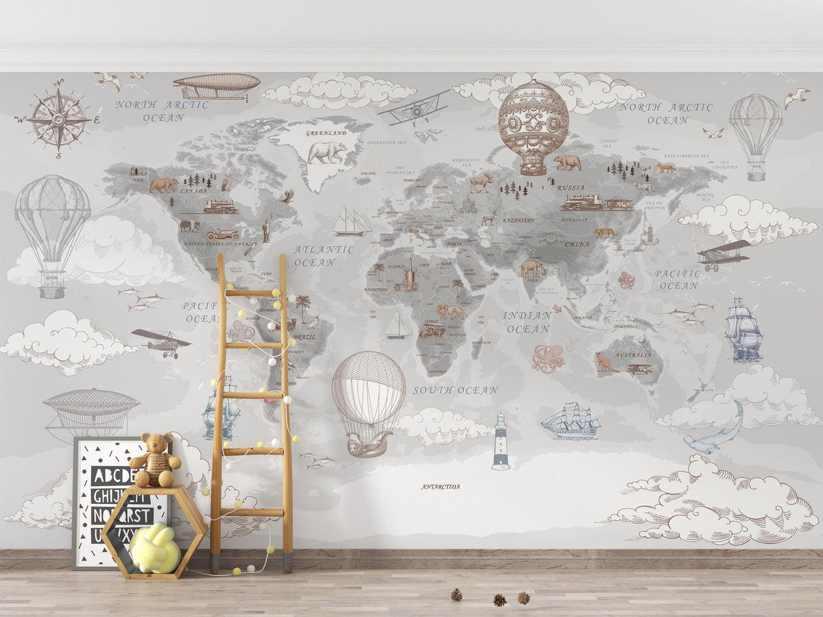 A children's room wall featuring a large vintage-style world map mural with illustrated hot air balloons, ships, airplanes, clouds, and small decorative elements in soft gray, beige, and muted blue tones. A wooden ladder, framed art, teddy bear, and small toys are placed on the floor in front of the mural.