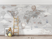 A children's room wall featuring a large vintage-style world map mural with illustrated hot air balloons, ships, airplanes, clouds, and small decorative elements in soft gray, beige, and muted blue tones. A wooden ladder, framed art, teddy bear, and small toys are placed on the floor in front of the mural.