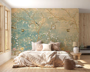 A styled bedroom featuring a large wall mural with a vintage-style floral and bird design in teal, beige, and gold tones. The room includes a bed with neutral-toned bedding, pillows, a small wooden side table with a lamp, a window on the left, and light wood flooring.