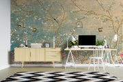 A styled home office interior featuring a large wall mural with a vintage-style floral branch pattern in teal, gold, pink, and brown tones. In front of the wall is a light wood sideboard, a white desk with a computer, white chairs, lamps, and a black-and-white chevron rug.
